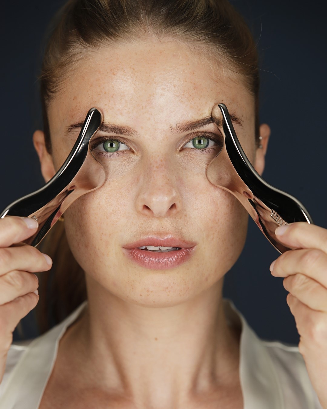 Eine Frau mit Sommersprossen verwendet ein silbernes Gua Sha-Werkzeug auf ihrer Stirn, um die Haut zu massieren und die Durchblutung zu fördern.