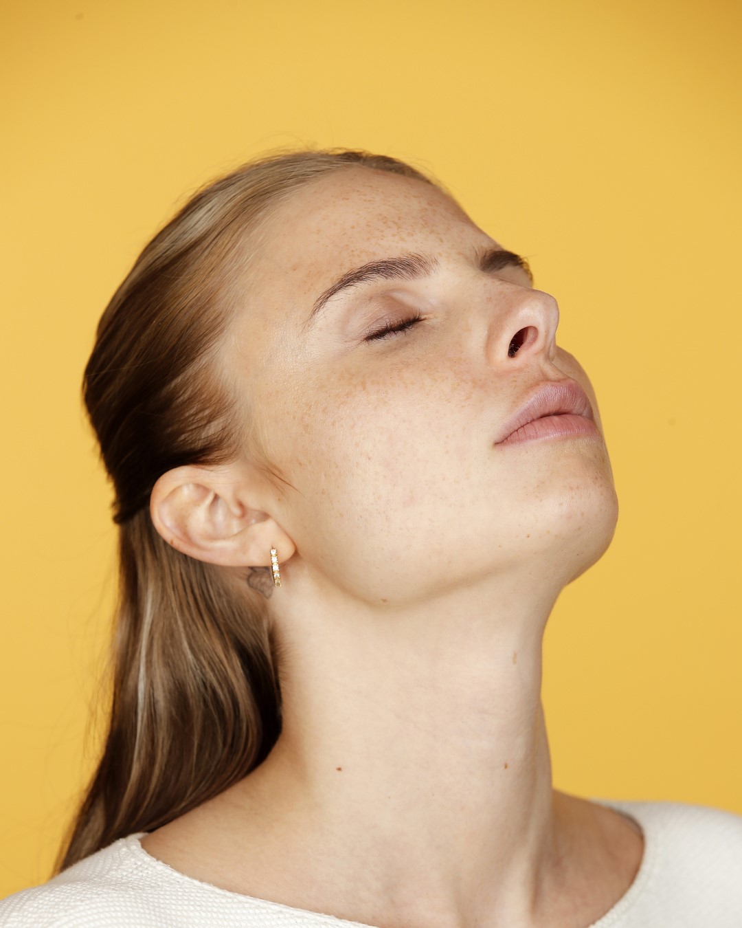 Eine Frau mit Sommersprossen und geschlossenen Augen, den Kopf leicht nach hinten geneigt, vor einem gelben Hintergrund.