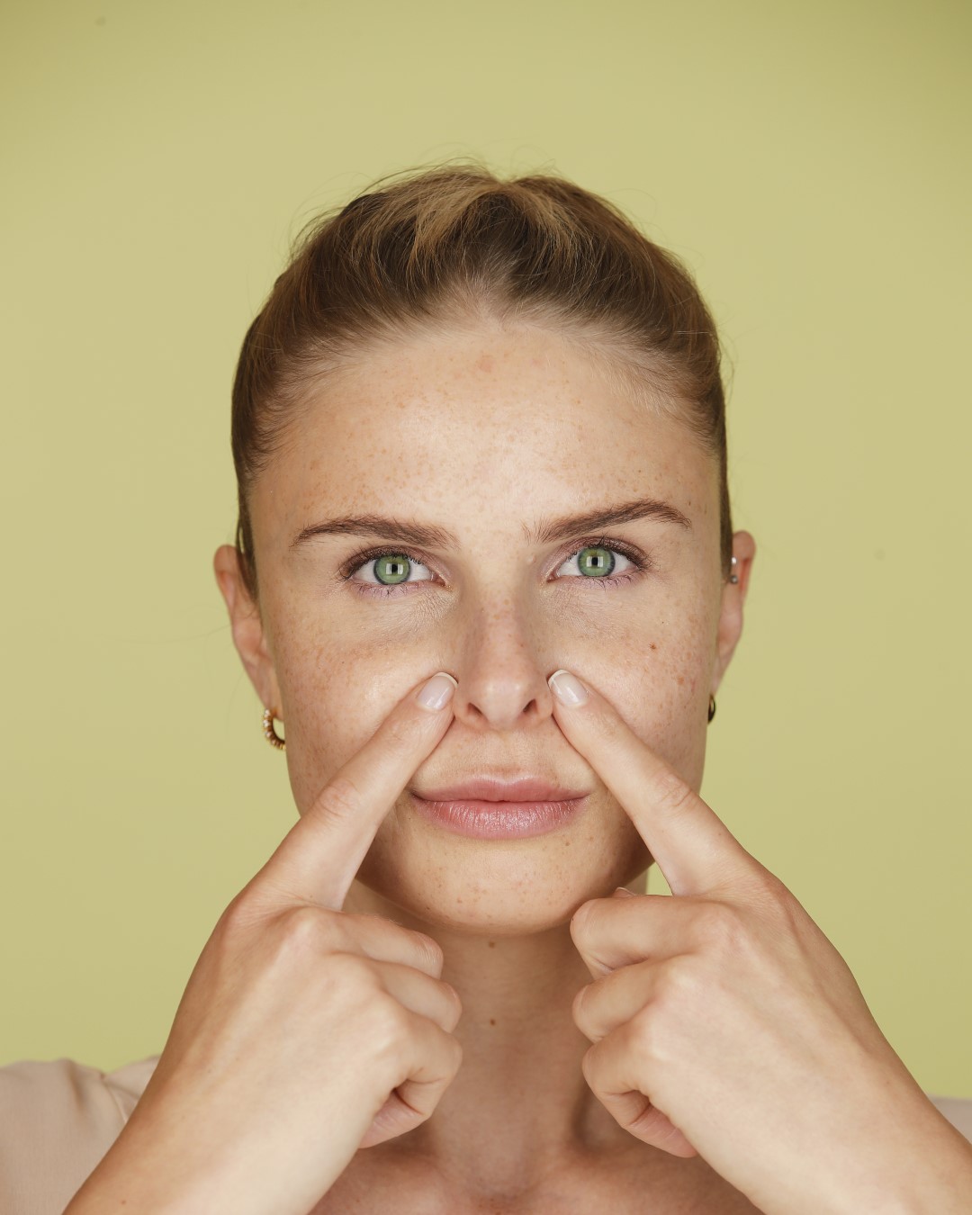Eine Frau mit grünen Augen und Sommersprossen drückt mit beiden Zeigefingern auf ihre Nasenspitze.