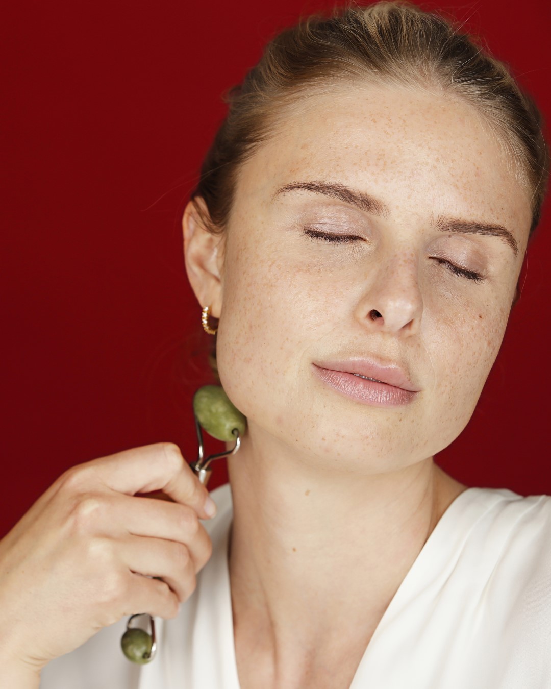 Eine Frau mit Sommersprossen genießt eine entspannende Gesichtsmassage mit einem grünen Jade-Roller, die Augen geschlossen und ein Gefühl des Wohlbefindens ausstrahlend.