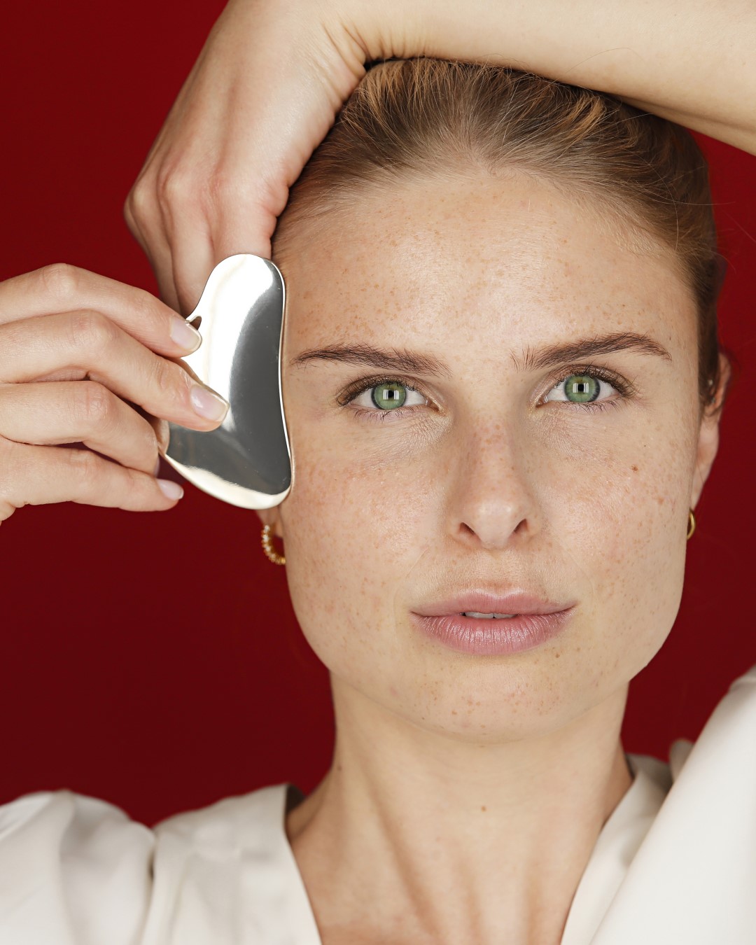 Eine Frau mit Sommersprossen und grünen Augen massiert ihr Gesicht mit einem silbernen Gua Sha Werkzeug vor einem roten Hintergrund.