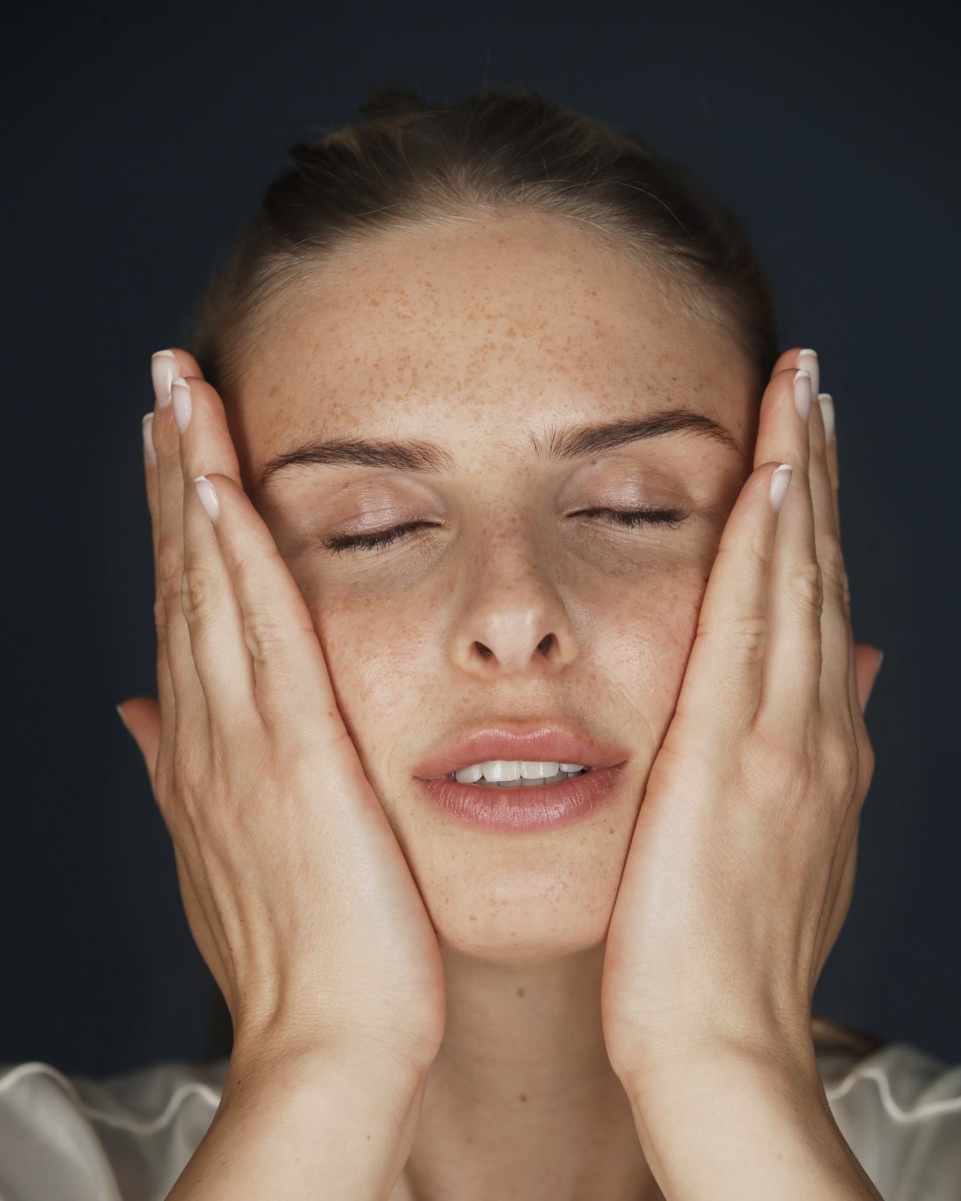 Eine Frau mit Sommersprossen berührt sanft ihr Gesicht mit geschlossenen Augen, um die Hautpflege zu genießen.