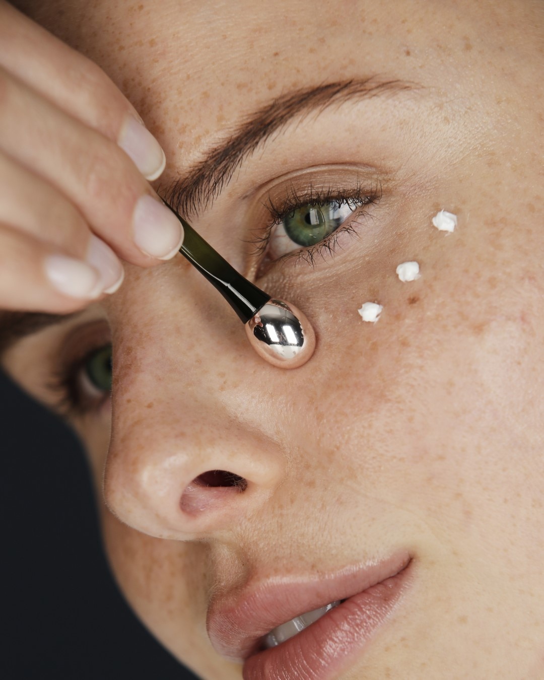 Nahaufnahme einer Frau, die mit einem silbernen Applikator eine weiße Creme unter dem Auge aufträgt, um die Haut zu pflegen.