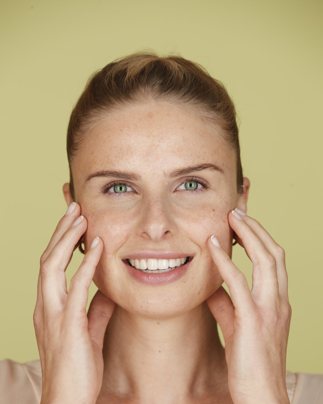 Nahaufnahme einer lächelnden Frau mit Sommersprossen, die ihre Wangen mit beiden Händen berührt, vor einem hellgelben Hintergrund.