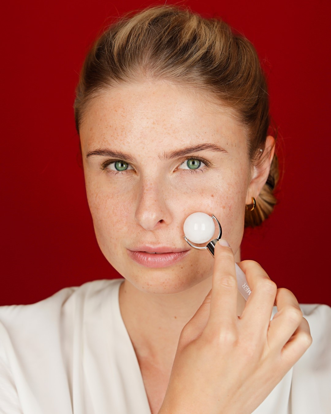 Eine Frau mit Sommersprossen massiert ihr Gesicht mit einem weißen Kugelroller vor einem roten Hintergrund, um die Hautpflege hervorzuheben.