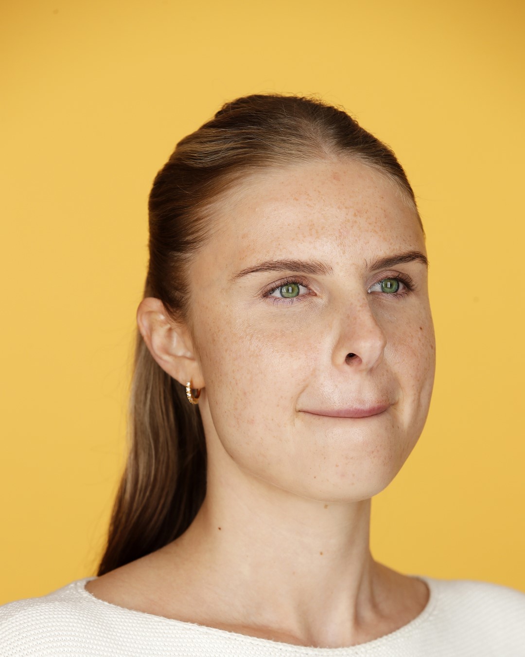 Porträt einer jungen Frau mit Sommersprossen, grünen Augen und blonden Haaren vor einem gelben Hintergrund.