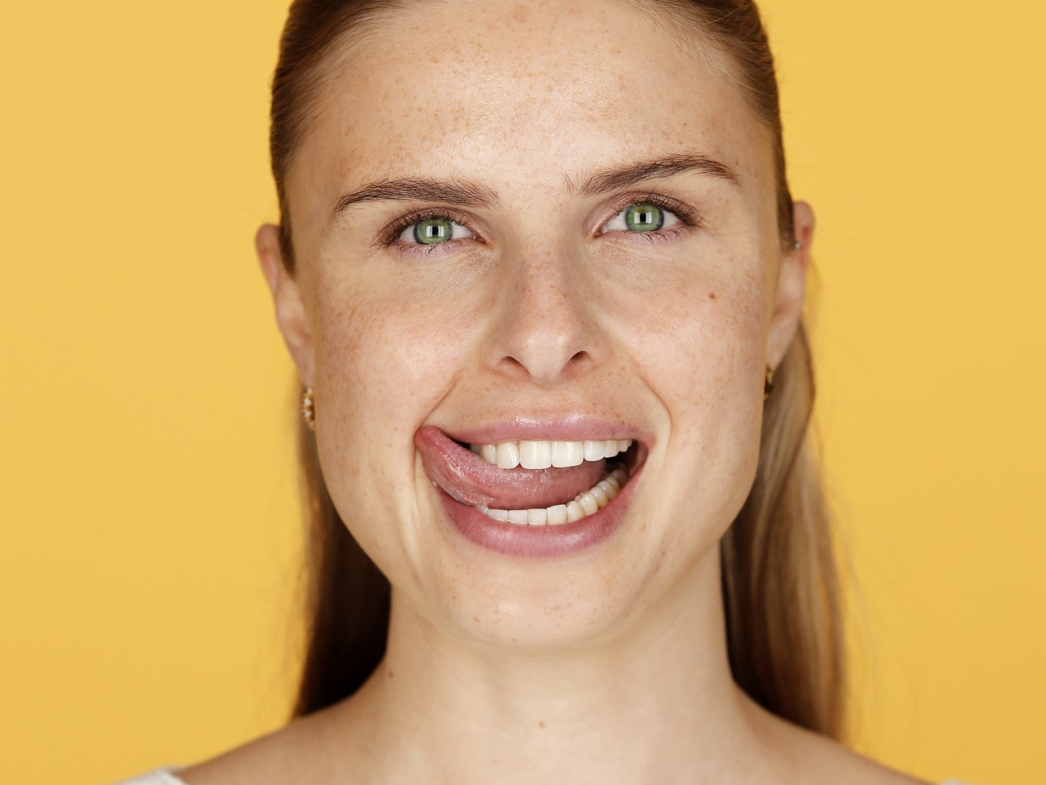 Nahaufnahme einer Frau mit Sommersprossen, die ihre Zunge herausstreckt und vor einem gelben Hintergrund lächelt.