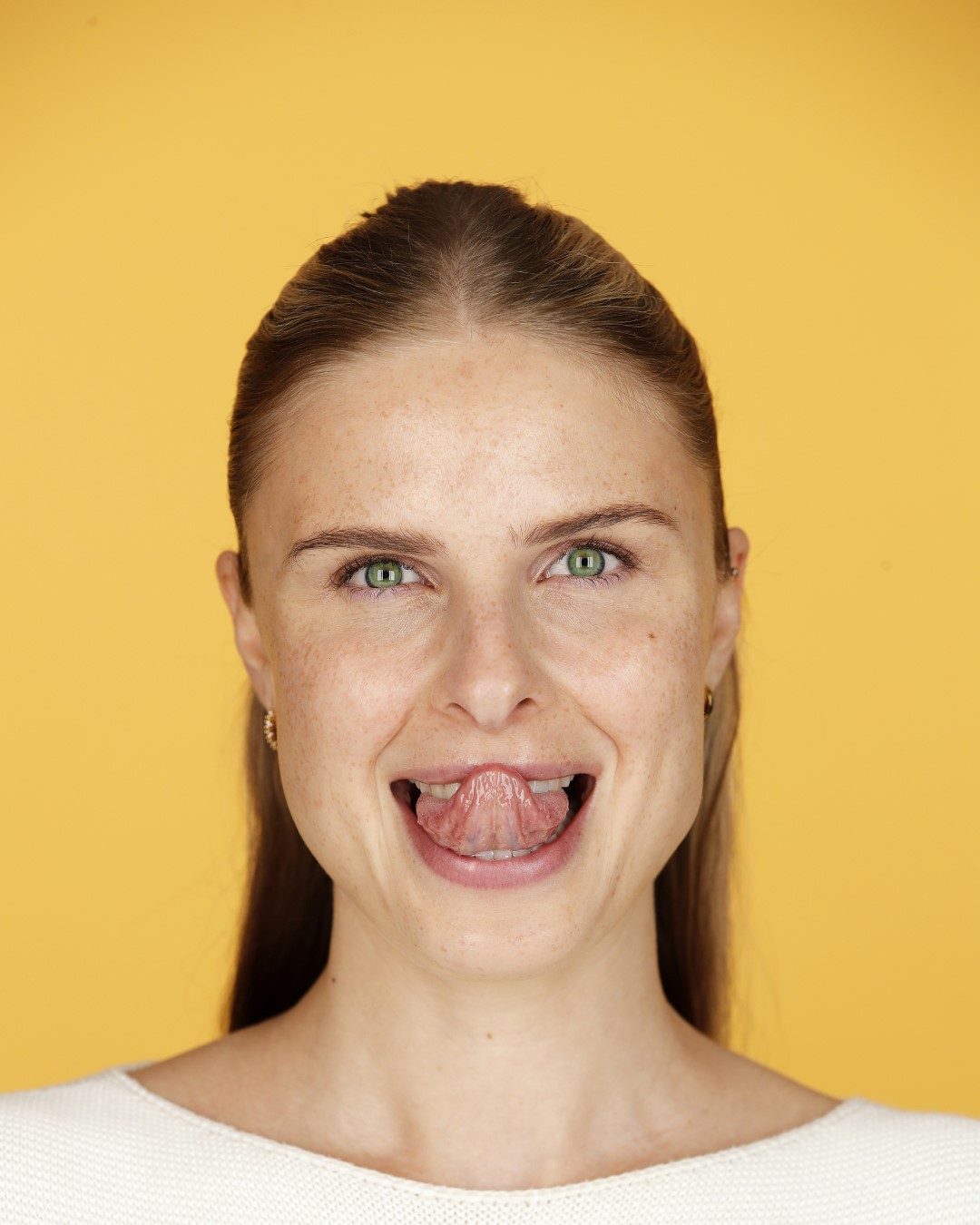 Eine Frau mit hellbraunem, zu einem Pferdeschwanz gebundenem Haar, Sommersprossen und grünen Augen streckt die Zunge heraus und lächelt vor einem gelben Hintergrund.