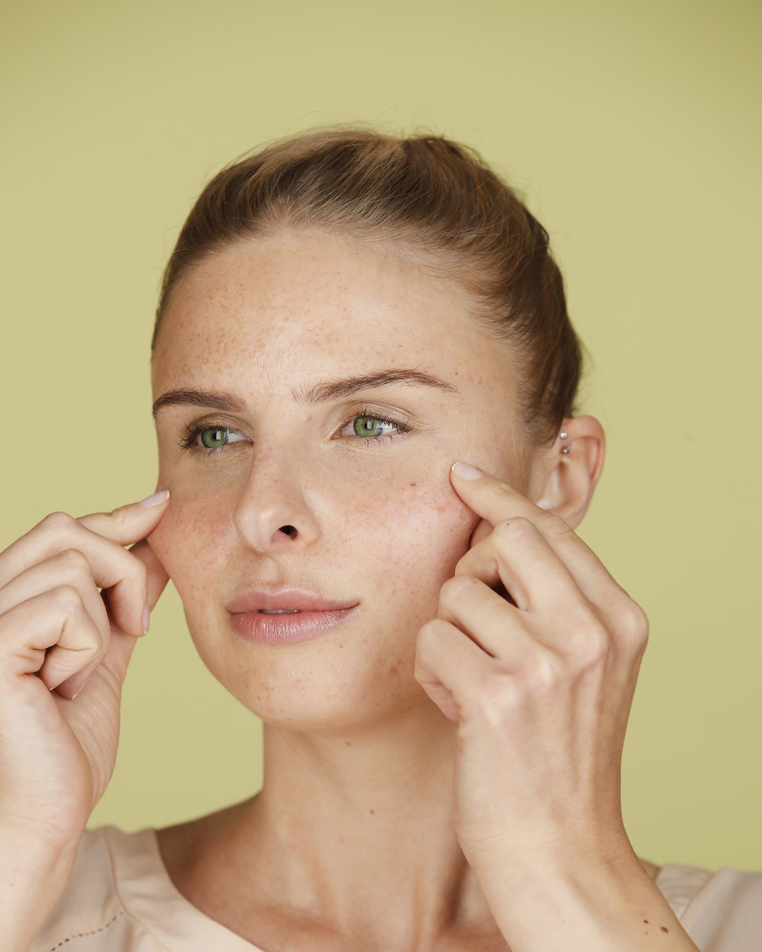 Eine Frau mit Sommersprossen berührt sanft ihre Wangen mit den Fingerspitzen, während sie mit einem leichten Lächeln zur Seite blickt.