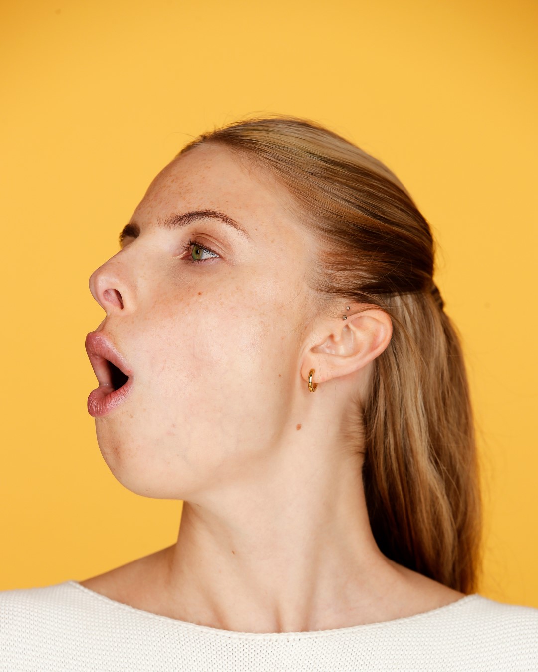 Profil einer jungen Frau mit blonden Haaren, die zu einem halben Zopf gebunden sind, und einem weißen Oberteil vor einem gelben Hintergrund, die ihren Mund weit geöffnet hat.