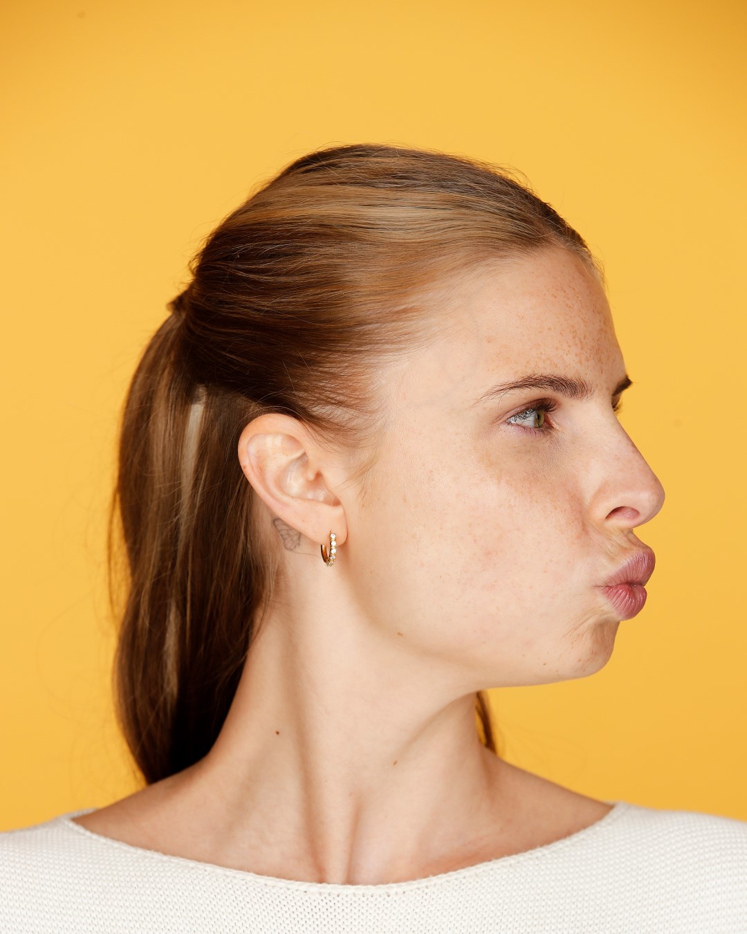 Profilansicht einer Frau mit Sommersprossen, die einen Kussmund macht und goldene Ohrringe vor einem gelben Hintergrund trägt.