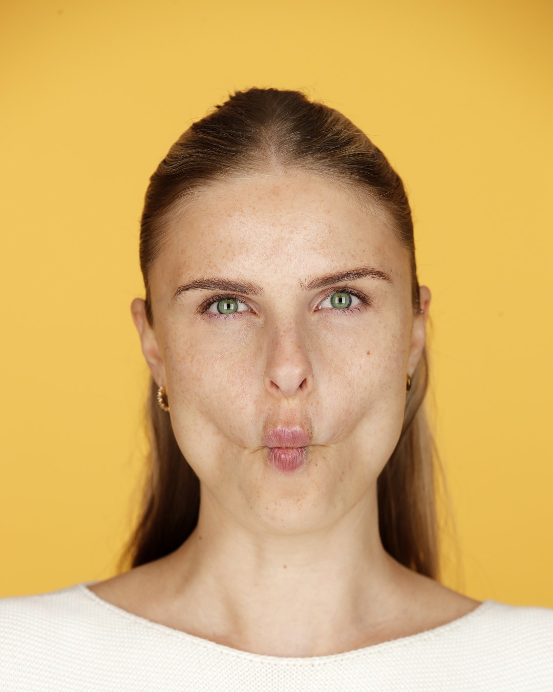 Porträt einer Frau mit hellbraunem Haar, grünen Augen und Sommersprossen, die einen Schmollmund zieht, vor einem gelben Hintergrund.