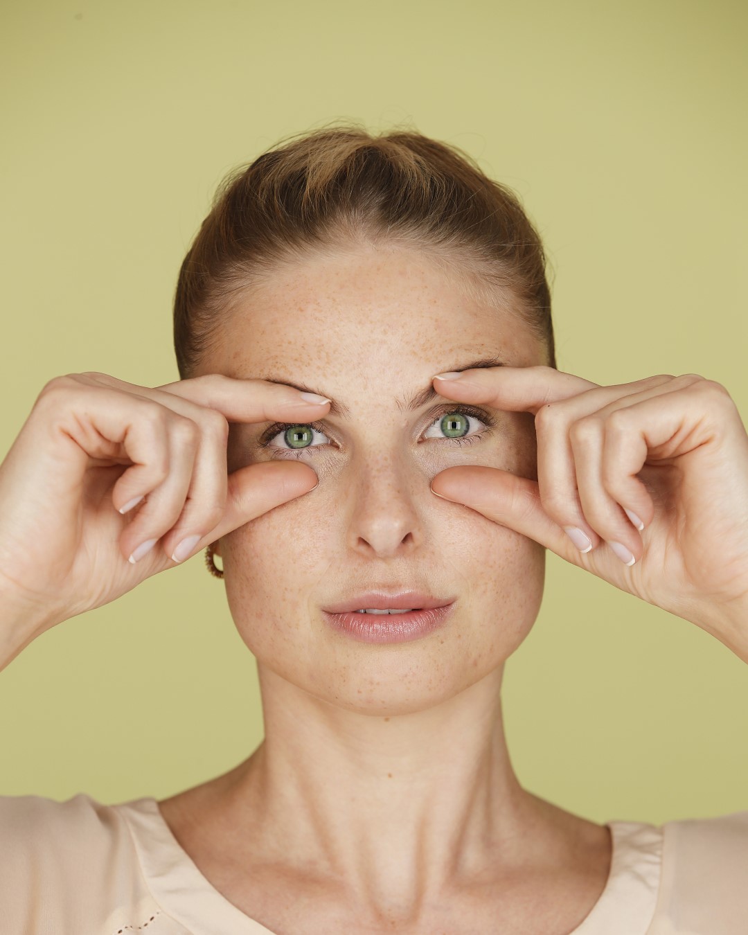 Eine Frau mit hellbraunem Haar und Sommersprossen hält ihre Zeigefinger an die Augenlider, um die Augenpartie zu straffen.