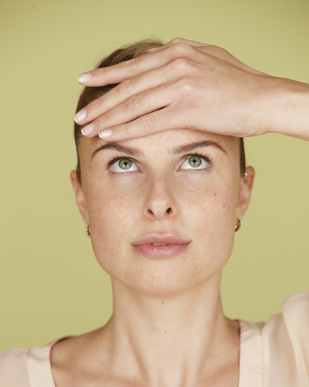 Eine Frau mit Sommersprossen legt ihre Hand auf ihre Stirn und blickt mit grünen Augen nach oben, während sie ein beiges Oberteil und goldene Ohrringe trägt.