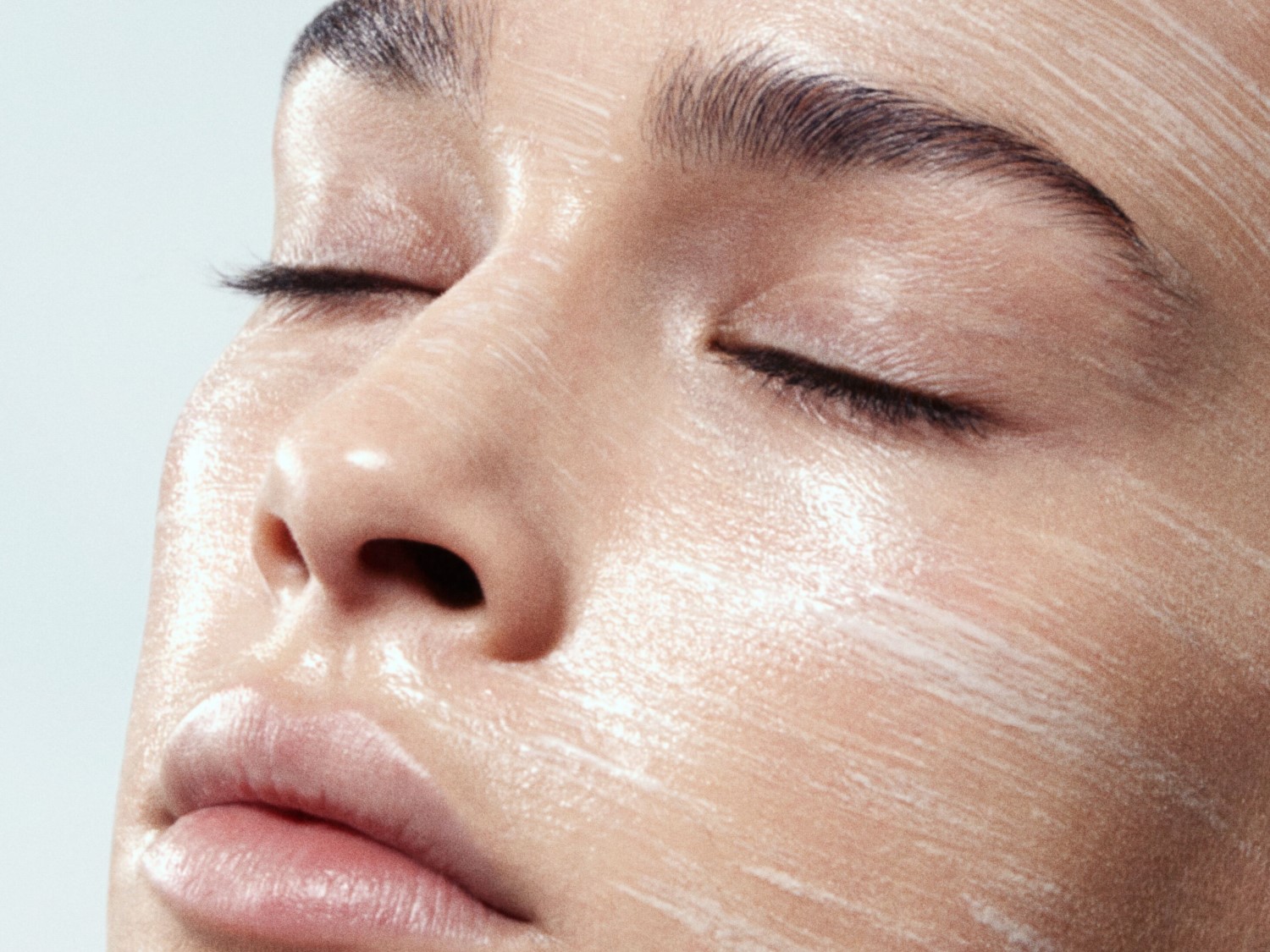 Primer plano del rostro de una mujer con los ojos cerrados y la piel cubierta con una crema blanca brillante, resaltando una tez hidratada y luminosa.