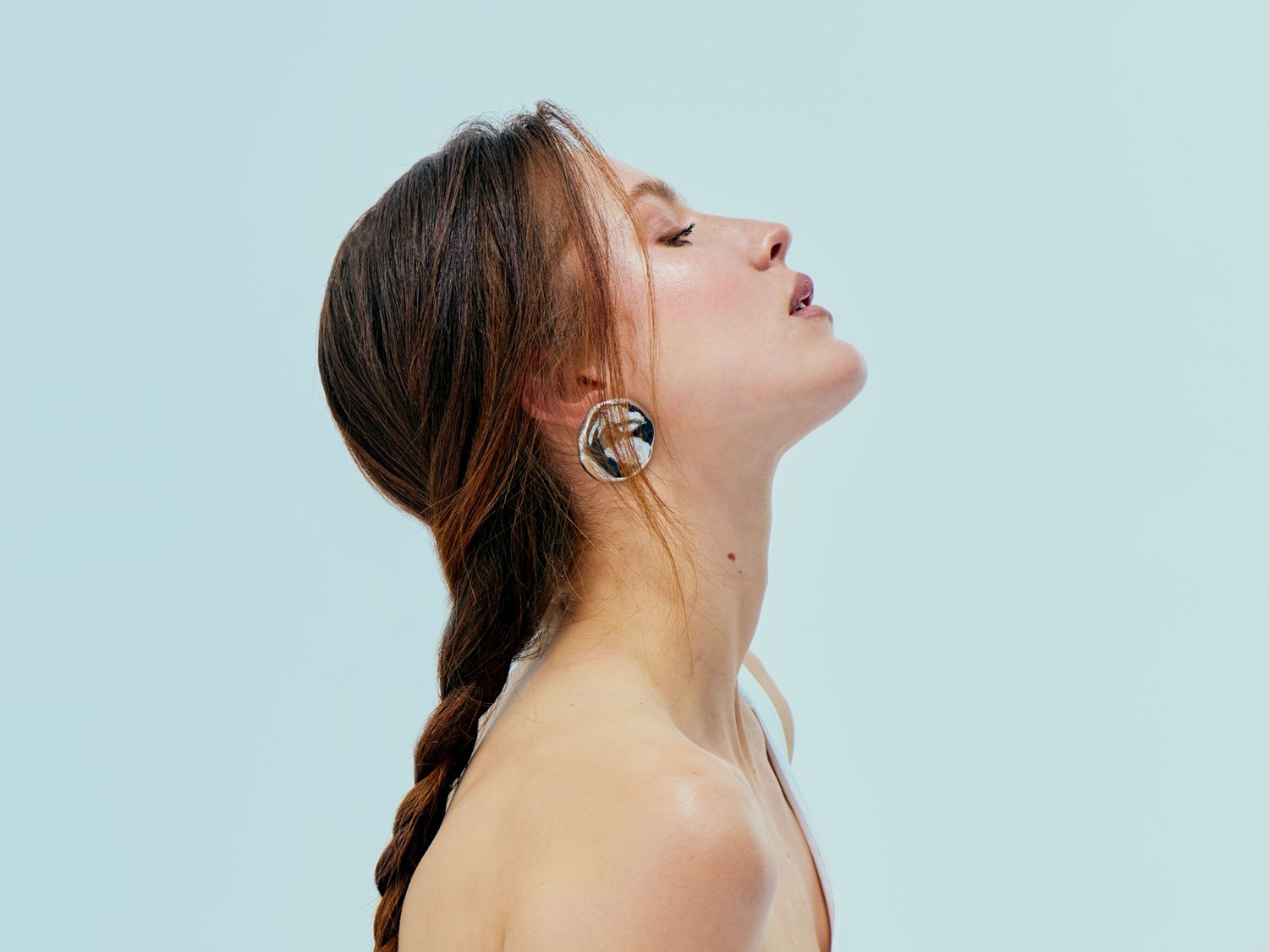Profil kobiety z brązowymi włosami zaplecionymi w warkocz, z głową odchyloną do tyłu, prezentującej okrągłe kolczyki na tle błękitnego nieba.