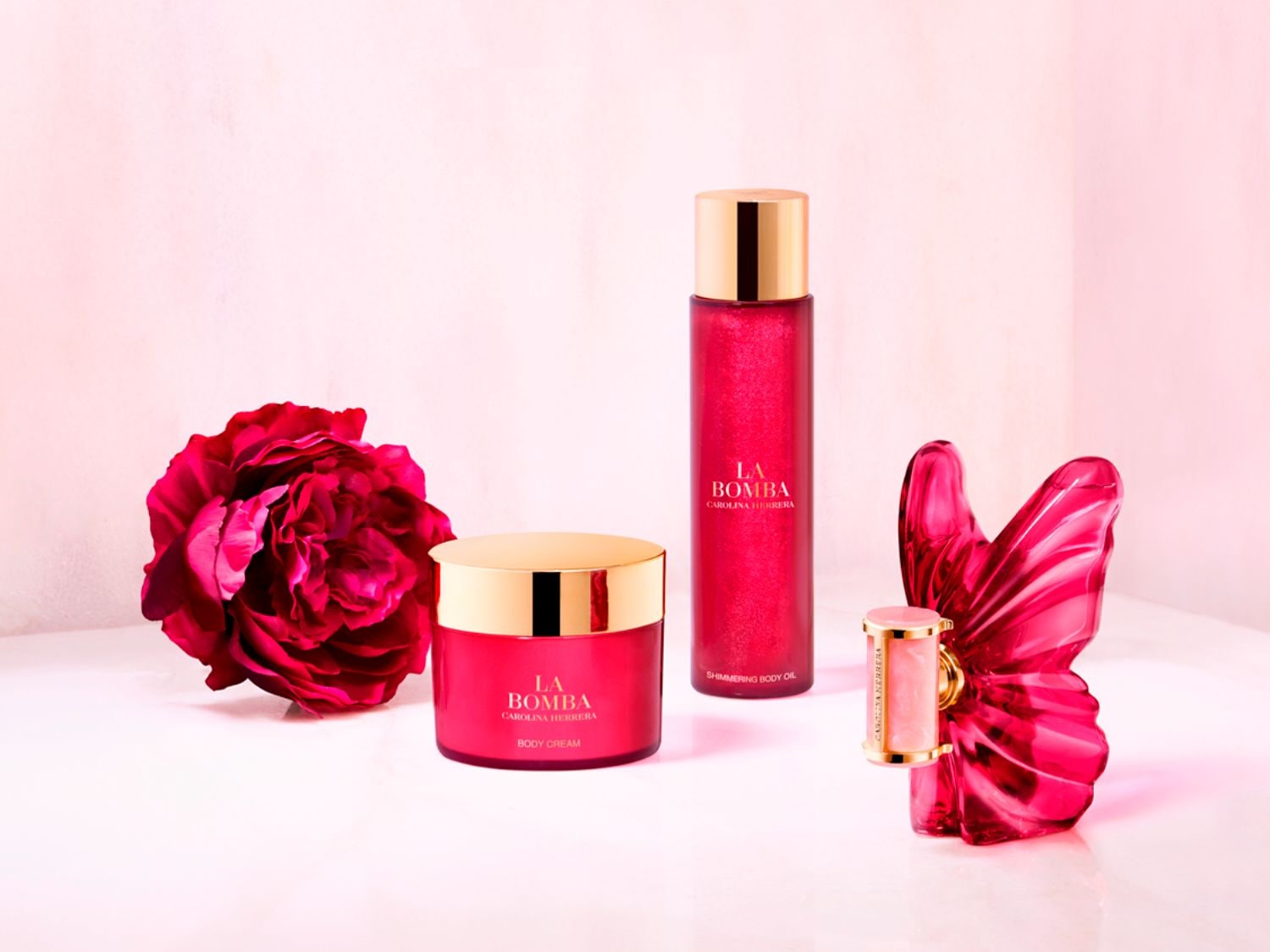 Telový krém La Bomba Carolina Herrera v ružovom balení so zlatým vrchnákom, vedľa neho je ružový kvet a v pozadí trblietavý telový olej.