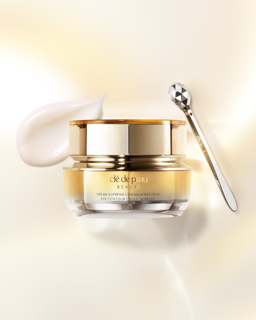Nahaufnahme von Clé de Peau Beauté Crème Supreme Contour des Yeux in einem goldenen Tiegel mit einem silbernen Applikator und einem Klecks weißer Creme.