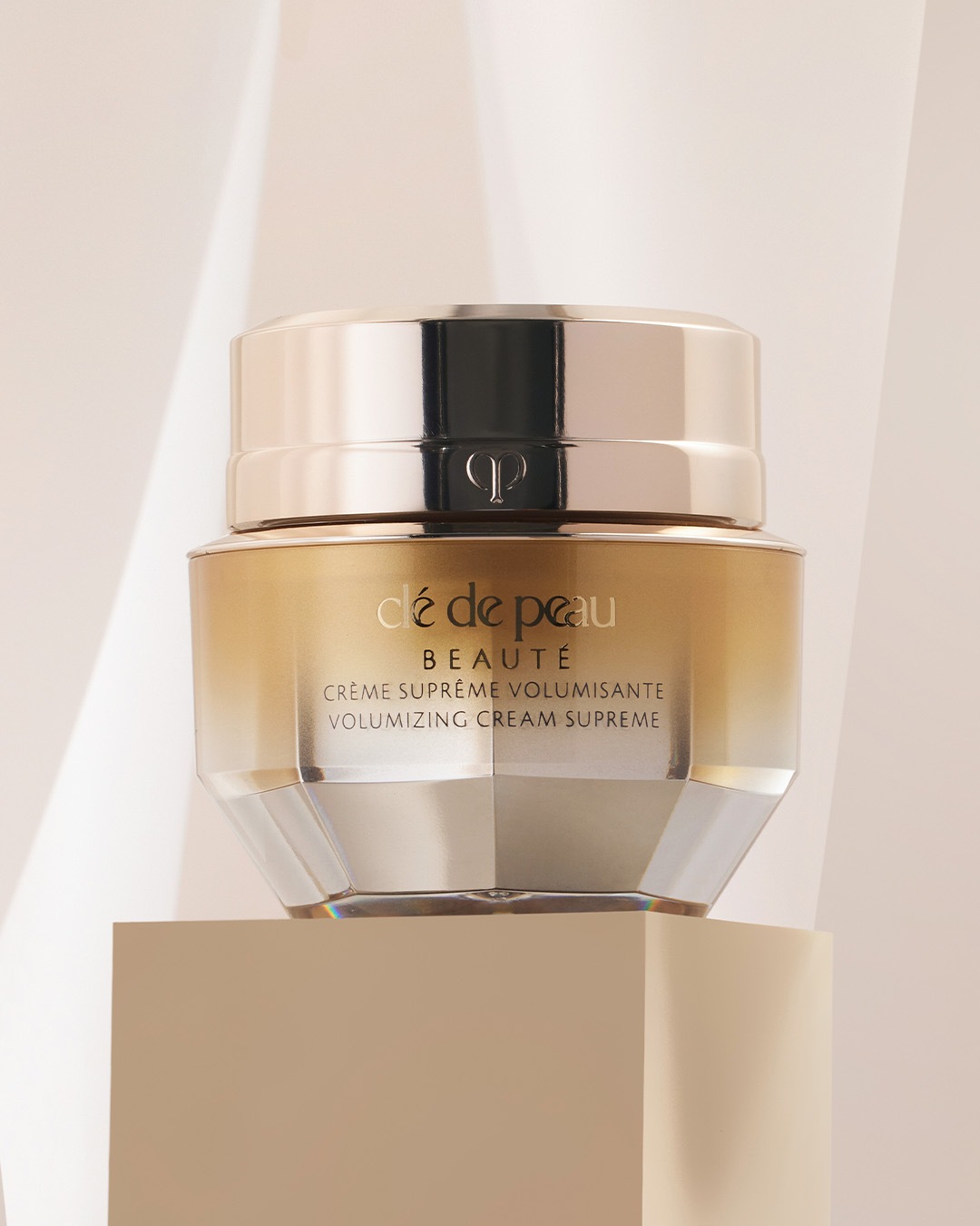 Nahaufnahme von Clé de Peau Beauté Volumizing Cream Supreme in einem goldenen Tiegel mit silbernem Deckel, der auf einem beigen Sockel steht.