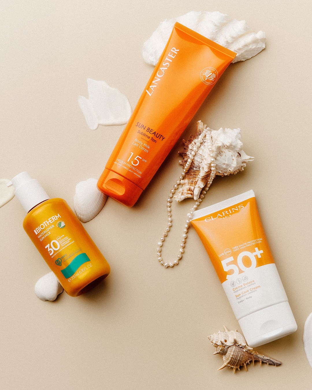 Tres protectores solares de Lancaster, Biotherm y Clarins con conchas marinas y un collar de perlas sobre un fondo beige.