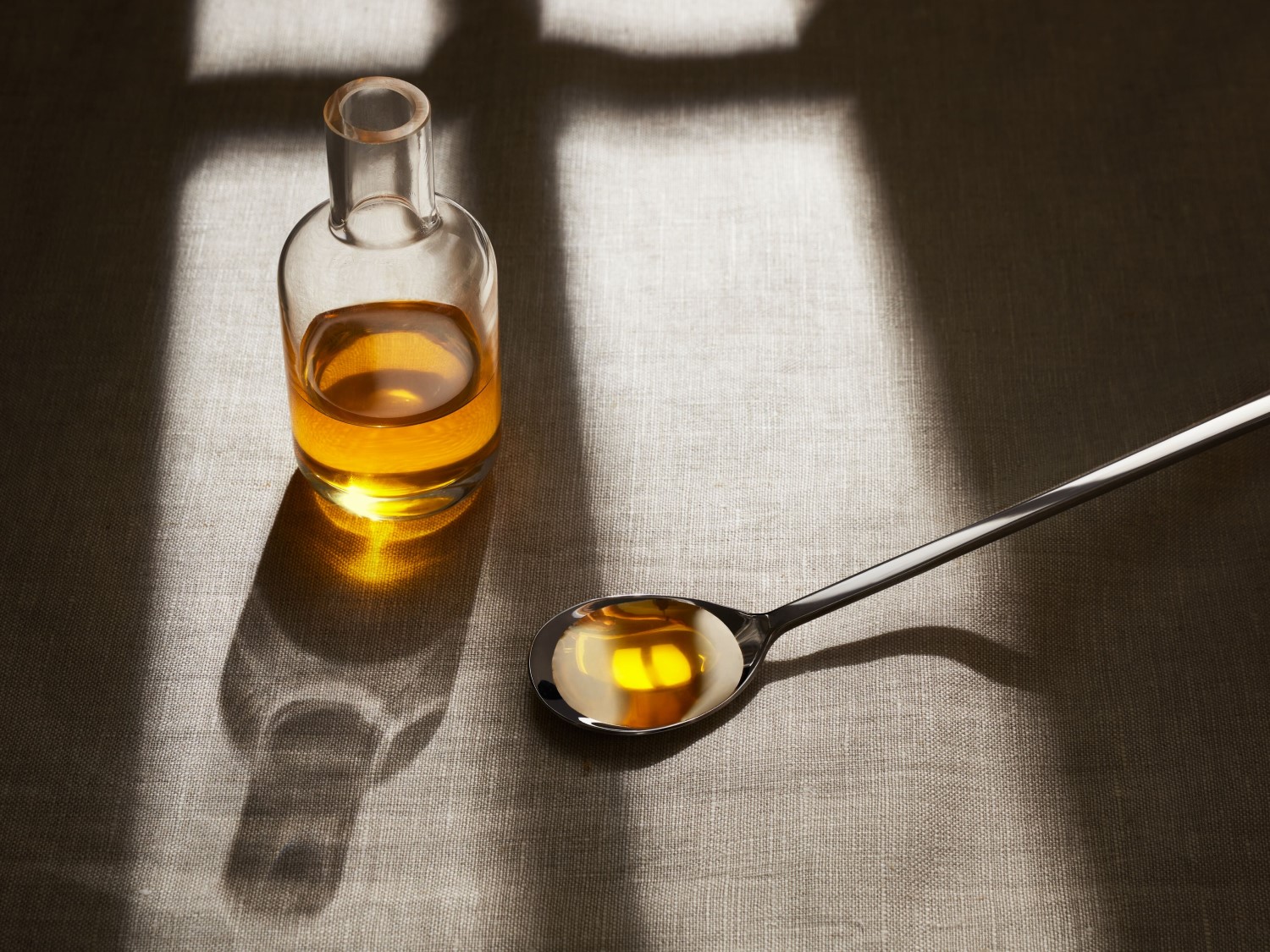 Una bottiglia di vetro trasparente riempita per metà con un liquido dorato, accanto a un cucchiaio d'argento anch'esso contenente il liquido, il tutto su un tessuto grezzo.