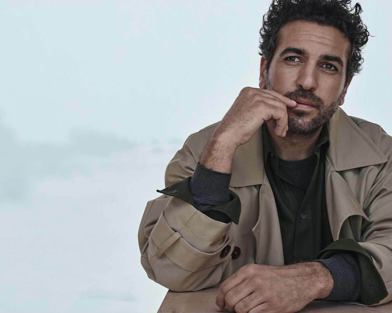 Ein Mann mit lockigem, dunklem Haar und Bart trägt einen beigen Trenchcoat über einem grünen Hemd und einem grauen Pullover, während er nachdenklich mit der Hand am Kinn sitzt.