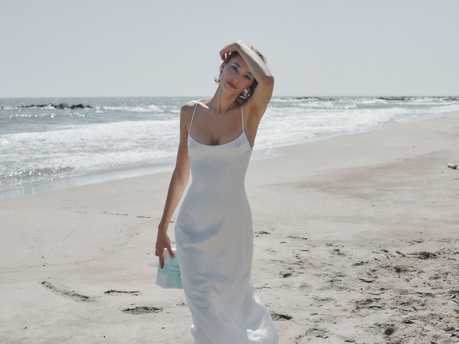 Eine Frau im weißen Kleid steht am Strand und hält eine hellblaue Schachtel in der Hand, während die Wellen sanft an den Ufern brechen.