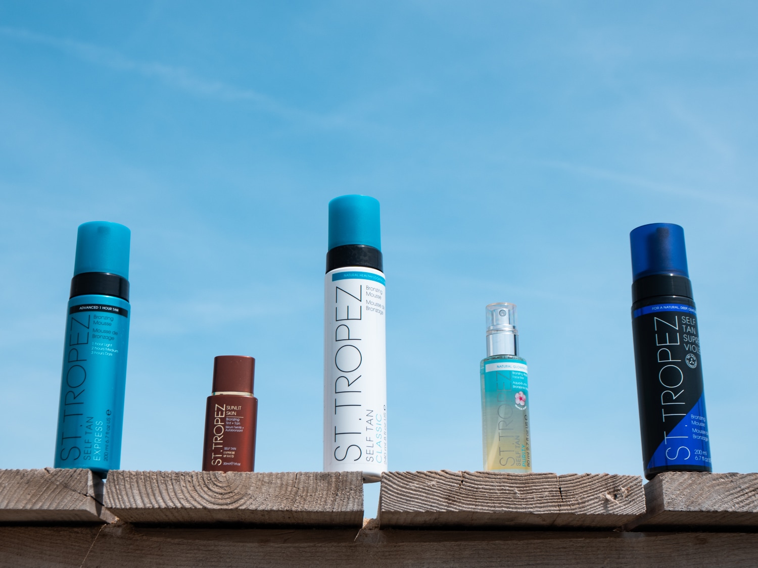 Różne produkty samoopalające St. Tropez są ustawione na drewnianej powierzchni na tle błękitnego nieba, w tym pianki brązujące i spraye w opakowaniach w odcieniach błękitu, brązu i bieli.