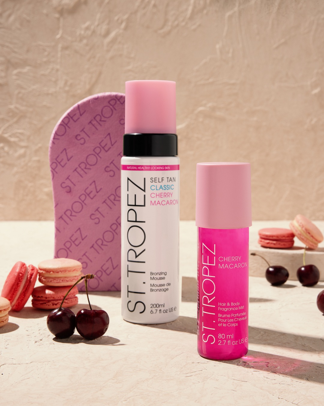 Biała butelka samoopalającego musu St. Tropez Classic Cherry Macaron z różową nakrętką, obok różowa mgiełka do włosów i ciała, wiśnie i różowe makaroniki.