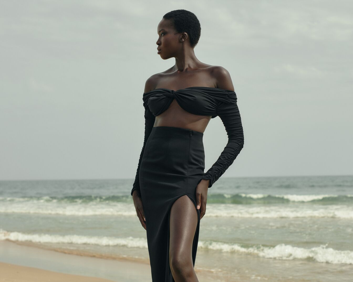 Eine dunkelhäutige Frau mit kurzen Haaren trägt ein schwarzes, schulterfreies Oberteil und einen Rock mit Schlitz vor dem Hintergrund eines Strandes.