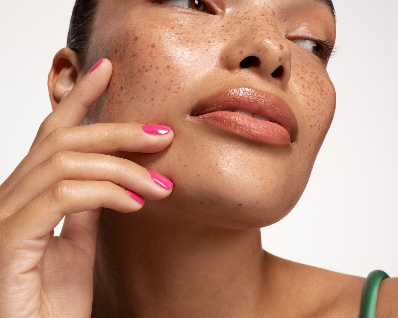 Primo piano di una donna con lentiggini, labbra color pesca lucide e unghie rosa acceso che tocca delicatamente la sua guancia.