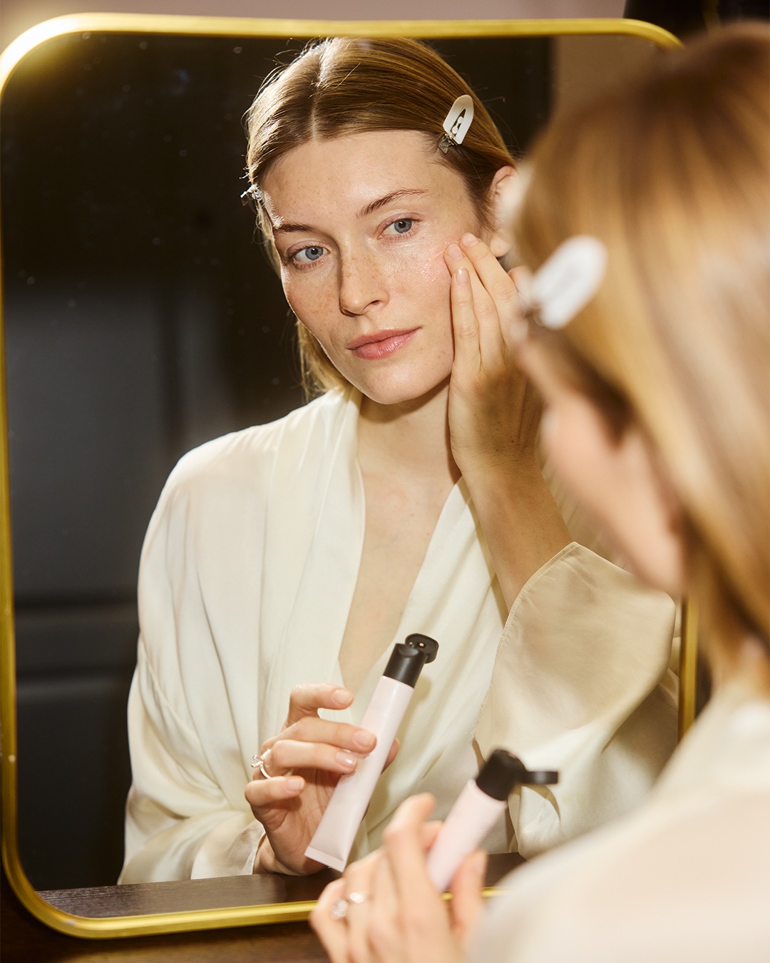 Een vrouw in een witte badjas staat voor een spiegel en brengt een roze tube met een zwarte dop aan op haar gezicht.
