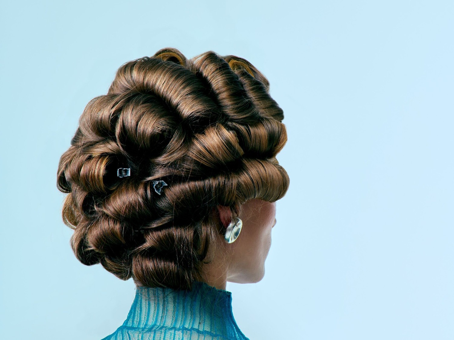 Nahaufnahme einer Frau mit aufwendiger, brauner Hochsteckfrisur, die kunstvoll zu Locken geformt und mit Haarspangen fixiert ist, dazu trägt sie ein blaues Oberteil und silberne Ohrringe.