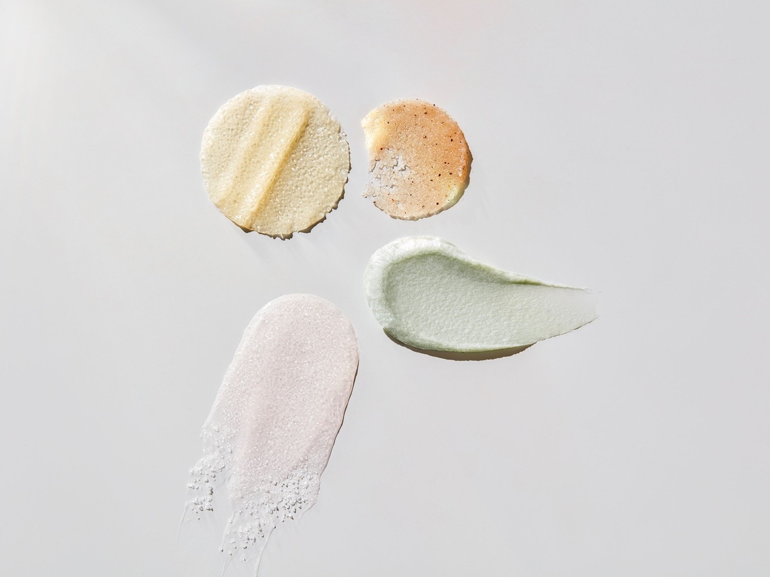 Verschiedene Texturen von Hautpflegeprodukten, darunter ein gelbes Peeling-Pad, ein orangefarbenes Peeling, eine grüne Creme und ein rosa Peeling, die die Vielfalt der verfügbaren Hautpflegeoptionen hervorheben.