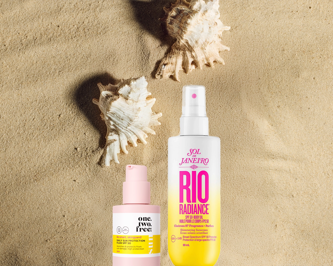 Sulla sabbia, conchiglie accanto a due flaconi di protezione solare: uno rosa "one. two. free!" e l'altro bianco e giallo "RIO Radiance SPF 50" di Sol de Janeiro.