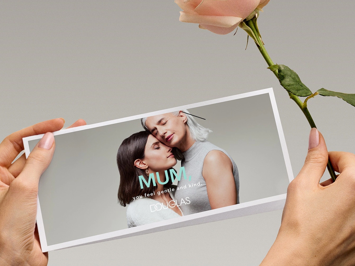 Una mano tiene una rosa color pesca, mentre l'altra tiene una foto di una madre e una figlia che si abbracciano con la scritta 'Mamma, ti senti gentile e premurosa' di DOUGLAS.
