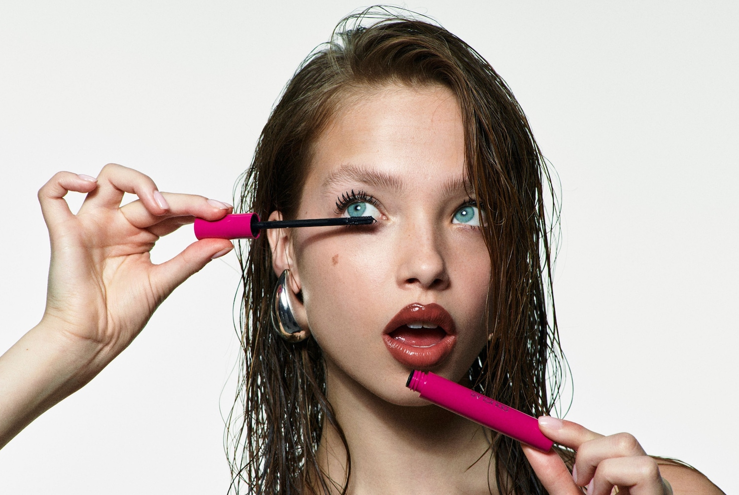 Une jeune femme aux yeux bleus applique du mascara rose vif sur ses cils, rehaussant son regard avec une touche de couleur audacieuse.