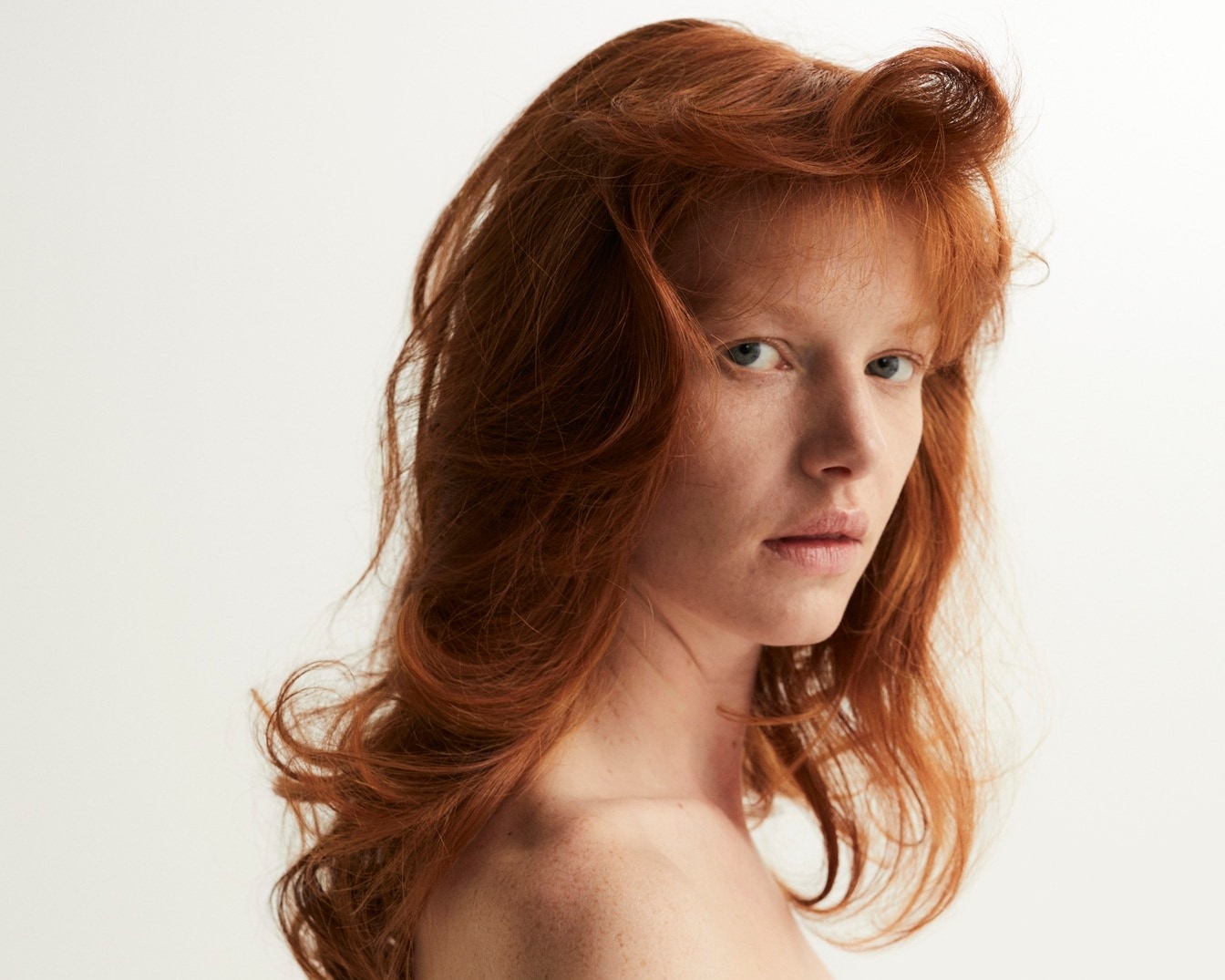 Portrait d'une jeune femme aux cheveux roux ondulés et aux yeux clairs, regardant fixement l'objectif avec une expression neutre.