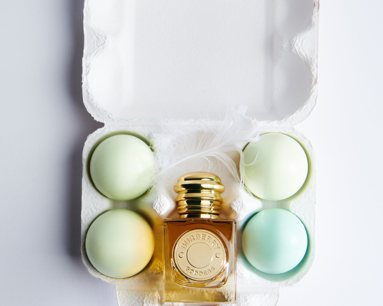 Dans une boîte à œufs, un flacon de parfum Burberry Goddess doré est entouré d'œufs pastel et d'une plume blanche, évoquant une ambiance printanière.