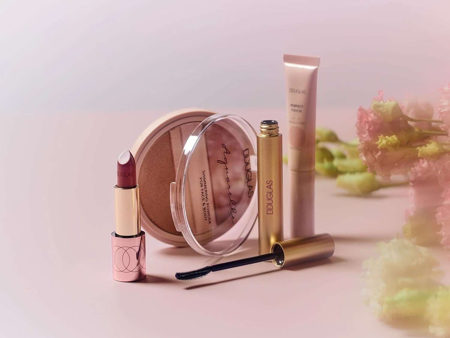 Eine Nahaufnahme von Douglas Make-up-Produkten, darunter ein Lippenstift mit burgunderroter Spitze, ein schimmerndes Puder für Gesicht und Körper, eine goldene Mascara und ein Perfect Touch Highlighter.