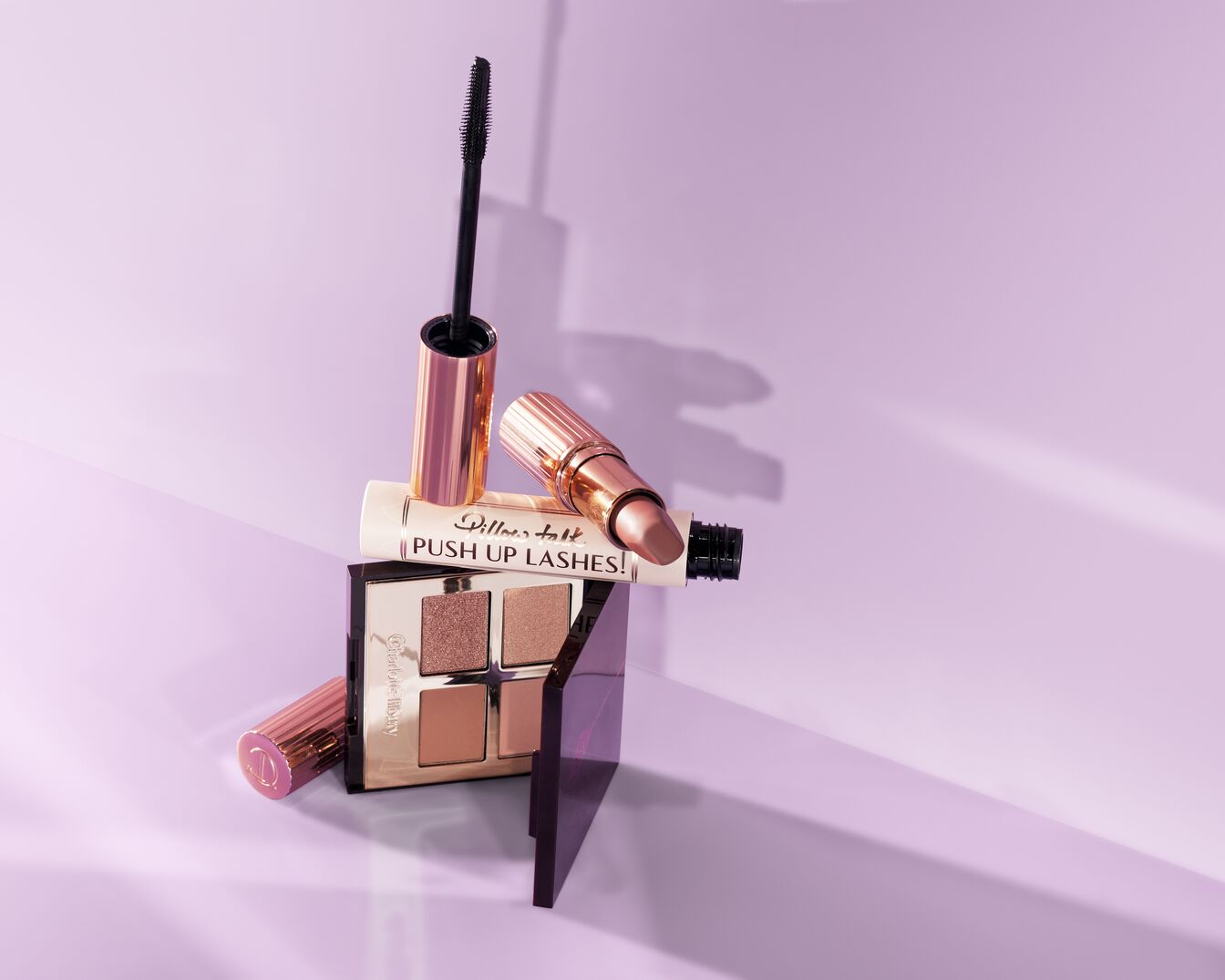 Nahaufnahme von Charlotte Tilbury Make-up Produkten, darunter Mascara, Lippenstift und Lidschattenpalette in Gold- und Rosatönen auf hellviolettem Hintergrund.