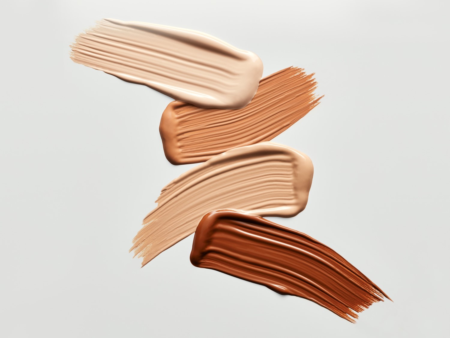 Muestras de maquillaje en varios tonos de piel, desde el beige claro hasta el marrón oscuro, dispuestas en trazos cremosos sobre un fondo neutro.