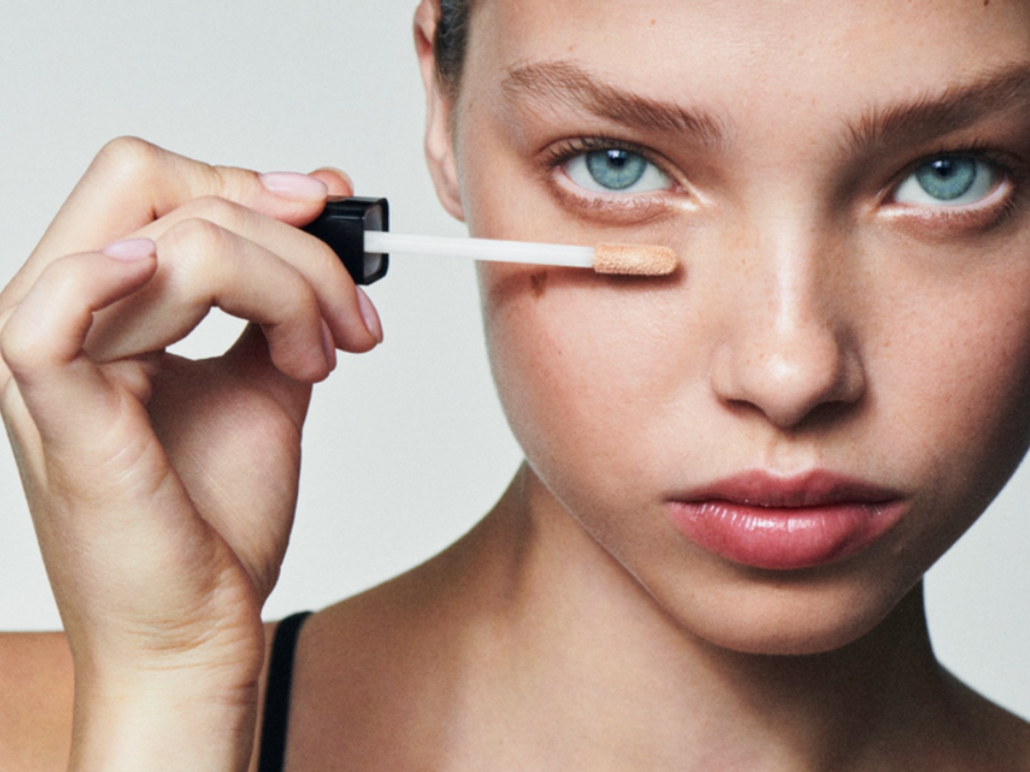 Primer plano de una mujer aplicando corrector beige con un aplicador blanco y negro para cubrir una mancha en su rostro, resaltando su piel clara y ojos azules.