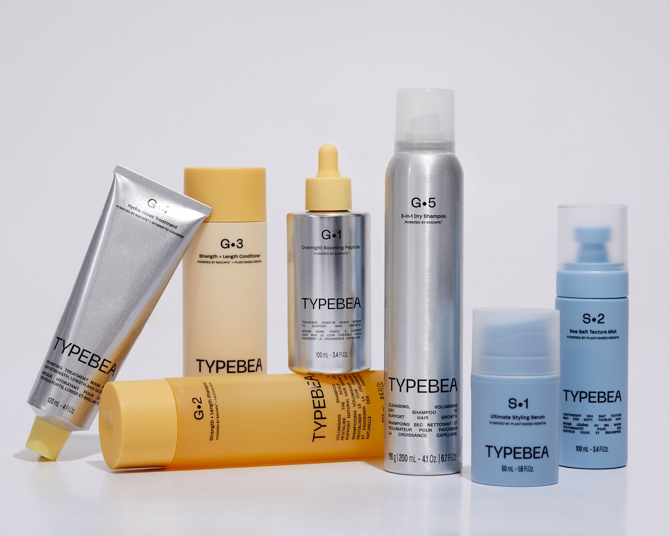 Een verzameling TYPEBEA haarverzorgingsproducten, waaronder hydraterende behandelingen, shampoos, conditioners, serums en droogshampoo, gepresenteerd in zilveren, gele en blauwe verpakkingen.