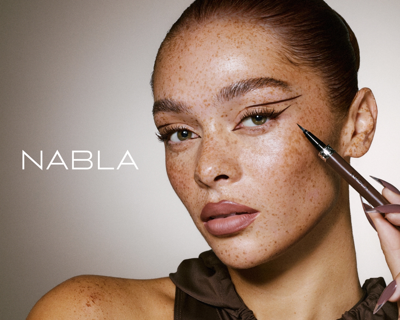 Una mujer de piel pecosa sostiene un delineador de ojos marrón de Nabla, mostrando un delineado gráfico y elegante que realza sus ojos verdes.