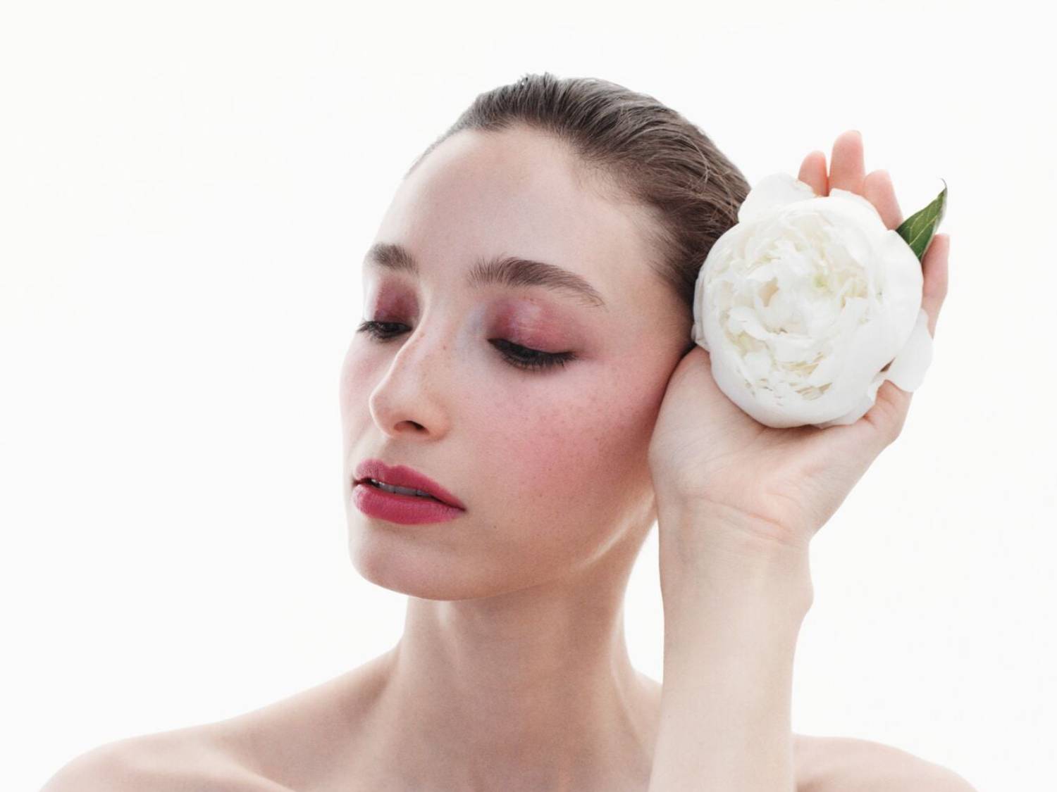 Una mujer sostiene una peonía blanca cerca de su rostro, con maquillaje rosa en los ojos y los labios, resaltando una belleza natural y delicada.