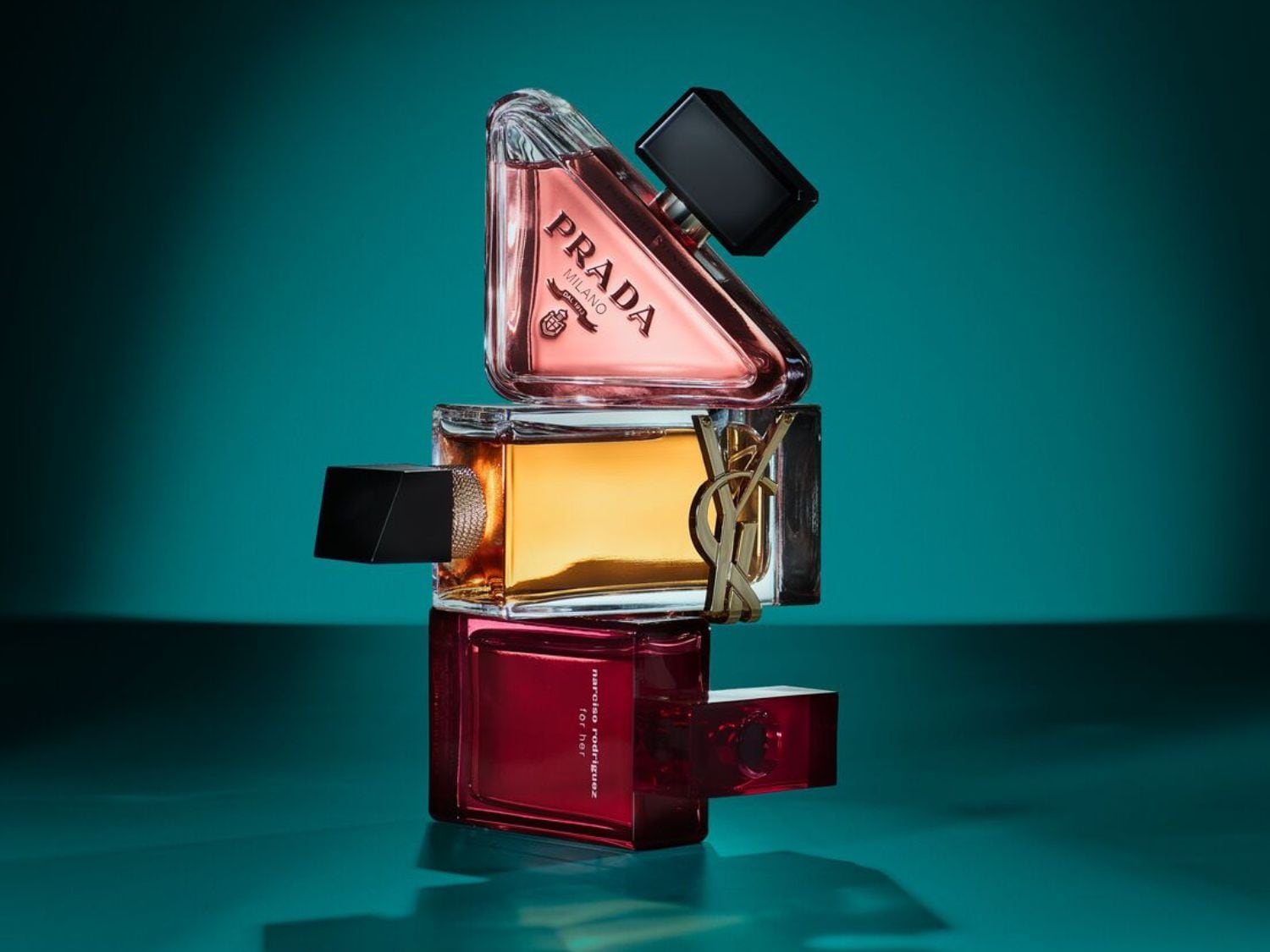 Na obrázku jsou tři flakony parfémů různých značek naskládané na sobě: Prada, Yves Saint Laurent a Narciso Rodriguez, všechny s výraznými tvary a barvami.