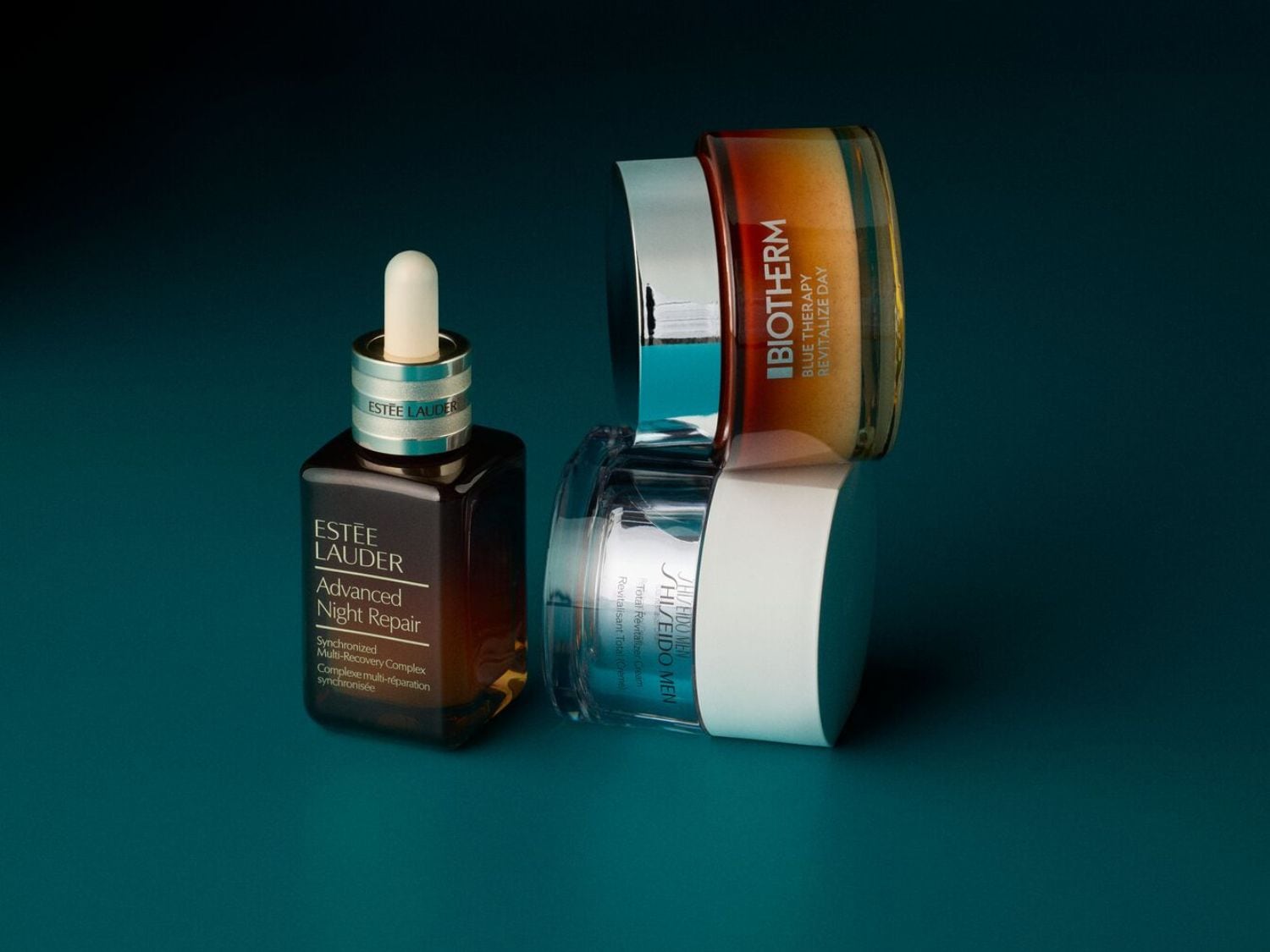 Hnědá lahvička Estée Lauder Advanced Night Repair se stříbrným hrdlem a bílým kapátkem stojí vedle stohu krémů Biotherm Blue Therapy Revitalize Day a Shiseido Men Total Revitalizer Cream.