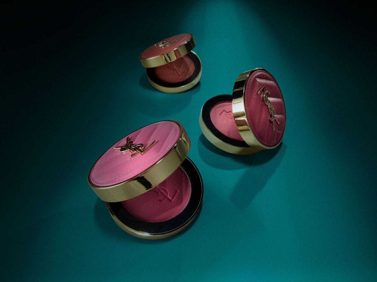 Tři otevřené pudřenky YSL s růžovým pudrem a zlatým logem na tyrkysovém pozadí.