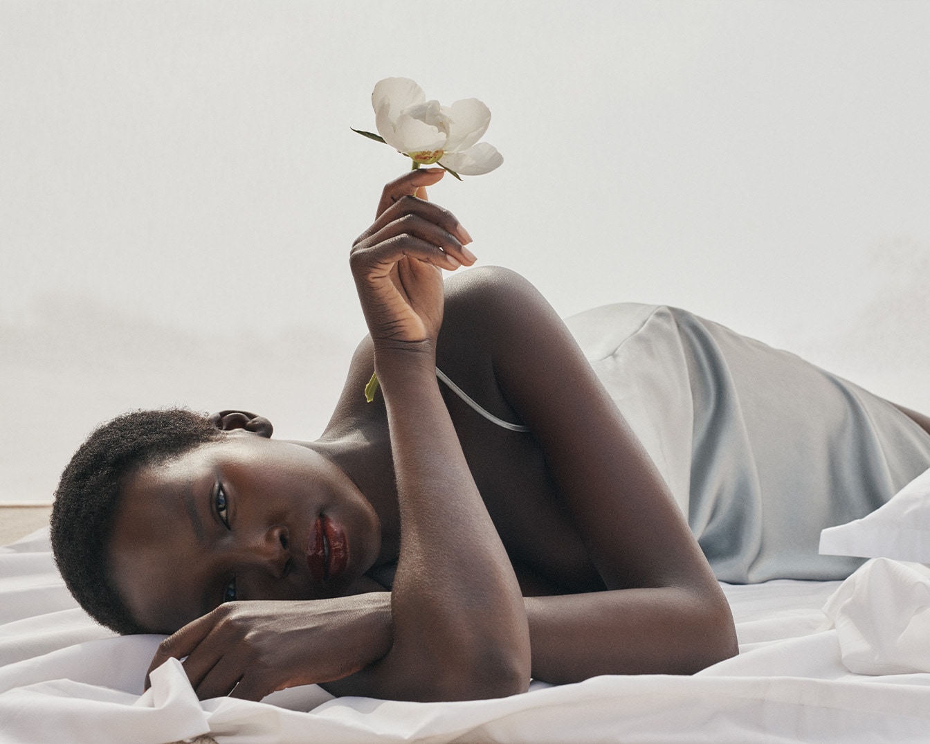 Eine dunkelhäutige Frau liegt auf einem weißen Tuch und hält eine weiße Blume in der Hand, ihr Blick ist sanft und einladend.